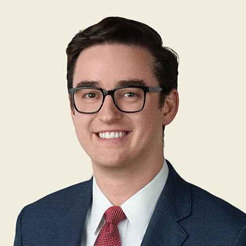 Zak Balasko; A young man with dark hair and glasses, wearing a navy suit, white shirt, and red tie, smiles confidently against a plain beige background. Professional tone.