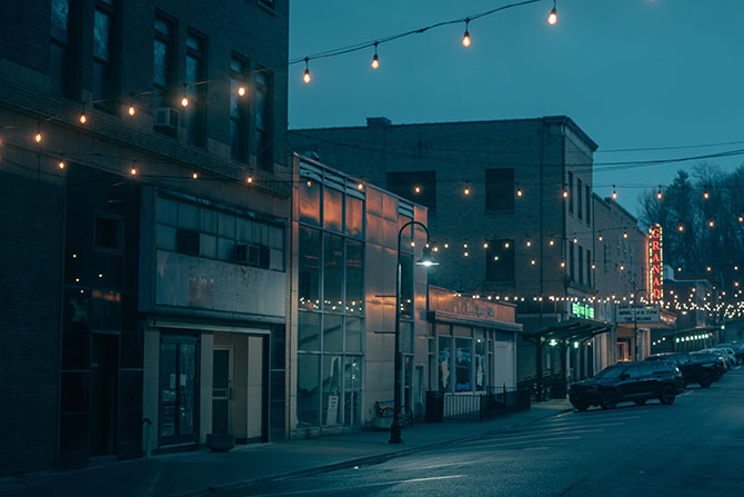 Investing in West Virginia’s Heritage; A dimly lit street at dusk, lined with quiet shops and twinkling string lights. A vintage theater sign glows red in the distance, creating a nostalgic ambiance.