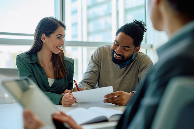 SBA Lending in a Changing Risk Environment; A man and a woman sit together in a bright office, smiling while discussing documents. A third person with a tablet listens nearby. Warm, collaborative atmosphere.