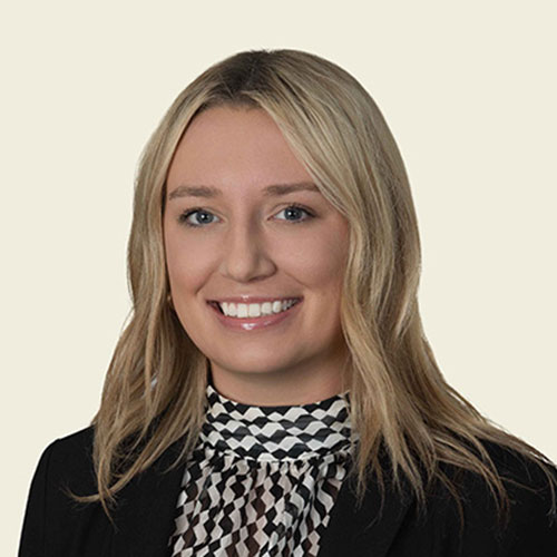Shelby Turley; A woman with blonde hair smiles, wearing a black blazer and a black-and-white patterned blouse, set against a plain cream background.